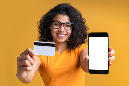 Close up isolated portrait of beautiful smiling african american girl showing mobile phone with empty white screen and blank credit card mock up directly at the camera. Copy space. Focus is on hands.の写真素材