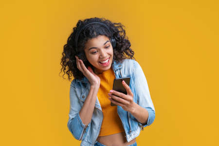 Cute smiling dark skinned girl in wireless headphones enjoying listening to music using application on her mobile phone.の写真素材