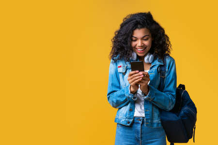Happy smiling casually dressed passenger with a backpack and bag browsing her mobile phone while waiting for her flight. Studio shot isolated over yellow background.の写真素材