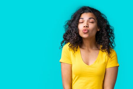 Studio shot of beautiful dark skinned girl in simple basic bright colored yellow t-shirt giving a kiss isolated over blue background.の写真素材
