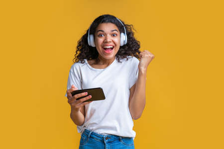 Studio shot of cute happy dark skinned girl wearing earphones, holding smartphone and looking at the camera smiling making winner gesture yes celebrating success.の写真素材