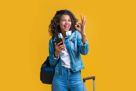 Happy cute casually dressed tourist girl with backpack and mobile phone winking and showing ok gesture. Isolated over yellow background.の写真素材