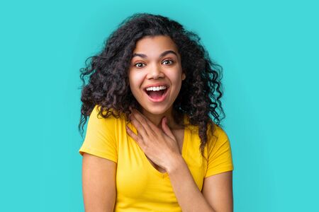 Portrait of beautiful young smiling dark skinned girl wearing bright colored yellow t-shirt isolated over blue background looking amazed with open-eyed face expression.の写真素材