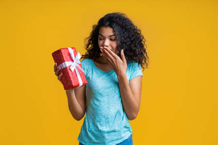 Portrait of astonished young african girl holding unexpected gift box decorated with satin ribbon covering her mouth with a hand in surpriseの写真素材