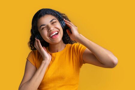 Studio portrait of cute smiling casually dressed dark skinned girl in wireless headphones enjoying listening to music and singing isolated over bright colored yellow backgroundの写真素材