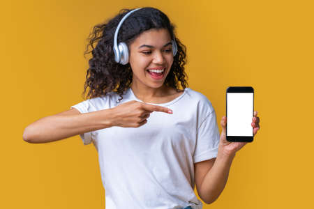 Cute smiling dark skinned girl in basic white t-shirt and wireless earphones showing blank screen of her smartphone at the camera. Mock up, copy space for your text, logo or interface.の写真素材