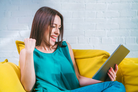 Attractive young woman sitting at home on yellow sofa with her tablet computer celebrating money win...