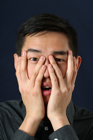 Astonished young Asian man looking at camera through his fingersの写真素材