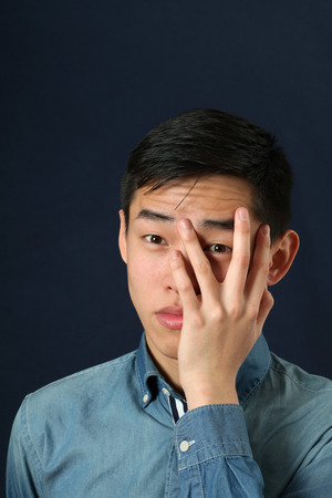 Displeased young Asian man looking at camera through his fingersの写真素材