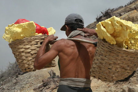 KAWAH IJEN, INDONESIA - AUGUST 8, 2011: Miner carries baskets with sulphur in the fumes of toxic volcanic gas from the sulphur mines in the crater of the active volcano of Kawah Ijen, East Java, Indonesia.のeditorial素材