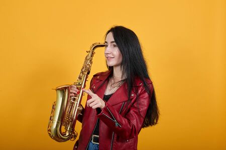 Portrait of amazed beautiful young woman in casual clothes looking, pointing index finger aside isolated on yellow orange wall background in studio, keeping saxophone. People lifestyle concept.の写真素材