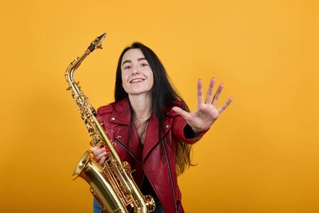 Portrait of crazy young woman in red casual clothes looking camera, keeping saxophone and smiling, standing with outstretched hands isolated on orange background. People lifestyle conceptの写真素材
