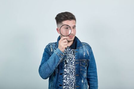 Young handsome man in fashion denim jacket holding a magnifying glass, looking on it, isolated on...