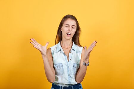 Caucasian young woman in blue denim shirt with closed eyes, spreads hands isolated on orange...