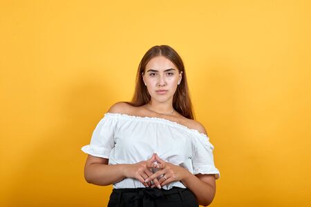 Strict young woman keeping hands together, looking at camera, serious girl isolated on orange background in studio in casual white shirt. People sincere emotions, lifestyle concept.の写真素材