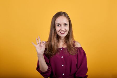 Young woman in burgundy bluse over isolated orange background showing sign okay with smile