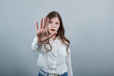 Attractive young woman in fashion white shirt doing stop, NO gesture, showing palm at camera isolated on gray background in studio. People sincere emotions, lifestyle concept.の写真素材