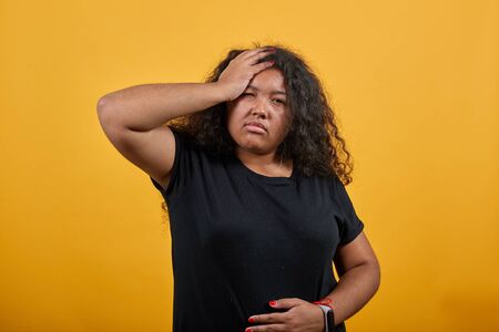 Sick afro-american young lady with overweight over isolated orange background wearing fashion black shirt keeping hand on head, headache, holding hand on stomach. People lifestyle concept.の写真素材