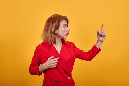 Attractive redhead caucasian young woman looking aside, showing finger at camera wearing fashion red jacket over isolated orange backgroundの写真素材