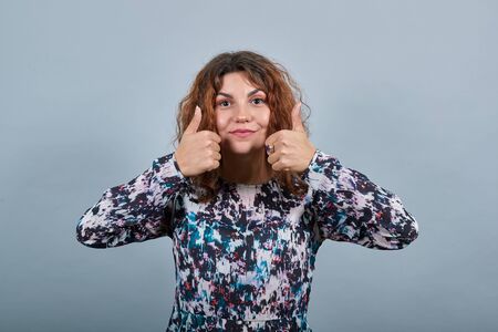 Attractive caucasian young woman over isolated gray background in fashion shirt keeping thumbs near...
