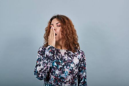 Attractive tired caucasian young woman covered mouth with hand, yawning over isolated gray background in fashion shirt. Lifestyle conceptの写真素材