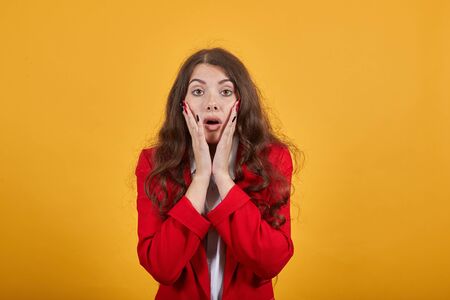 Surprised woman in fashion white shirt and red jacket keeping mouth opened, holding hands on cheeks, looking confused isolated on orange background in studio. People sincere emotions, lifestyle.の写真素材