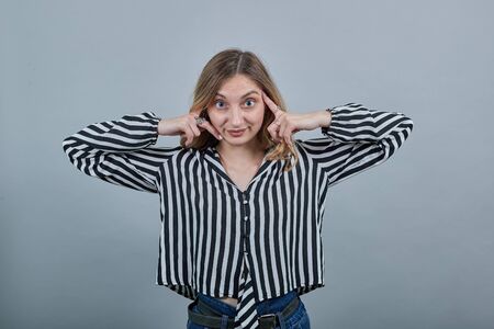 Shocked caucasian blonde woman in fashion black and white striped shirt looking at camera, keeping fingers on pulse isolated on gray background in studio. People sincere emotions, lifestyle concept.の写真素材