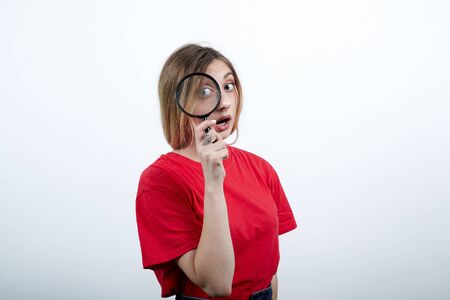 Serious caucasian woman keeping magnifier glass, looking on it wearing fashion red shirt isolated on white background in studio. People sincere emotions, lifestyle concept.の写真素材