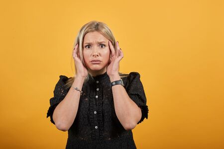 Young blonde girl in black dress on yellow background scared woman is afraid holds her hands to face