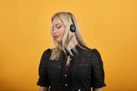 Young blonde girl in black dress on yellow background happy woman with headphones listening to music