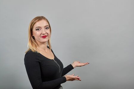 Young blonde girl in black jacket on grey background happy woman smiles and points the direction with her index finger, demonstrates somethingの写真素材