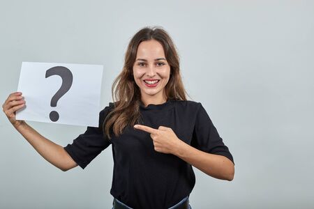 Cute Young Brunette Woman In Black T-Shirt, Blue Jeans With Belt On Gray Background, Happy Girl Smiling Holding Piece Of Paper With Question Mark In Hand And Pointing Index Finger In The Directionの写真素材