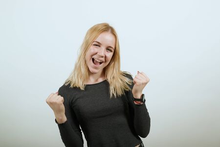 Young Blonde Woman In Black Sweater With Stylish Watch On White Background, Happy Girl Smiles Clenched Her Hands Into Fists And Raised Them Up. The Concept Of Successful, Purposeful Peopleの写真素材