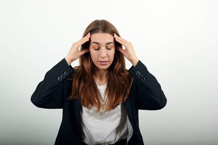 Headache grimacing pain holds the back of her neck indicating location. Fatigue during workaholism labor. Young attractive woman with brown hair in a light t-shirt and black jacket on white backgroundの写真素材