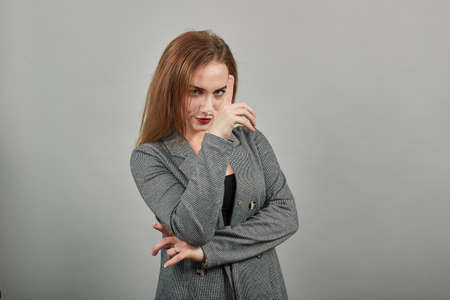 Showing loser sign, displaying dud gesture, alpha. Two fingers 2, disgust on face, negative human emotion facial expression. Young attractive woman, dressed gray jacket, with green eyes, light brownの写真素材