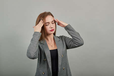 Headache grimacing pain holds the back of neck indicating location. Fatigue during workaholism labor. Young attractive woman, dressed gray jacket, with green eyes, light brown hair, backgroundの写真素材