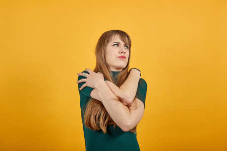 Hugging herself to warm while feeling cold arms crossed fear expression on face loneliness and depression. Being terrified with unknown sound. Young attractive woman, dressed green shirt blonde hairの写真素材