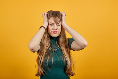 Headache grimacing pain holds the back of neck indicating location. Fatigue during workaholism labor. Young attractive woman, dressed green shirt blonde hair, yellow backgroundの写真素材