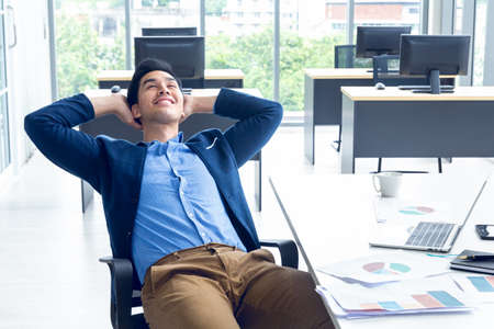 Top view of young businessman sitting in a modern office. He has a feel happy and smile about the result of business profits is positive. On his table have a computer laptop tablet pen paper graph and a coffee cupの写真素材