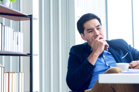 A young businessman sitting in a modern office. He has a feel sleepy because  hard work so tired weary fatigued and exhausted. On his table have a computer laptop tablet pen paper graph.の写真素材