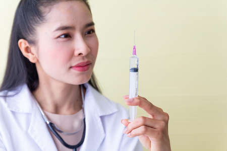 The female Asian doctor wearing a white robe sitting in a chair with a stethoscope on the neck be a serious face and preparing a syringe for patients.の写真素材