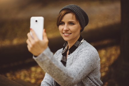 Portrait of smiling and happy young millenial woman sitting alone in the forest, taking selfie and posting to social media with smartphone. Broadband mobile internet connectionの写真素材