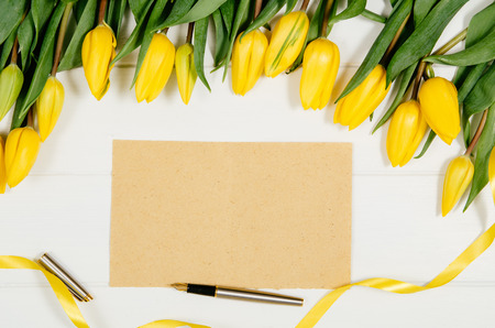 Background with yellow tulips, blank notepad on white wooden table top view. Creative womans workspace concept with copy space. Product photograph taken from above with frame compositionの写真素材