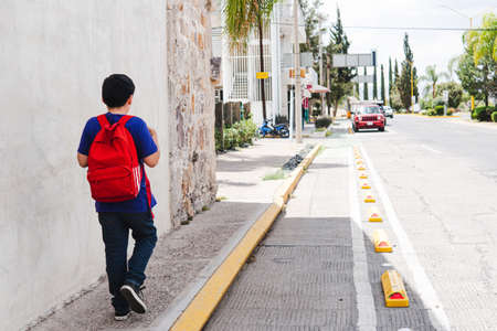 Black-haired boy with a red backpack and wearing a purple shirt walking to his school