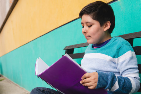 A boy in a colorful sweater sitting on a bench with a green wall behind him reading a purple book...