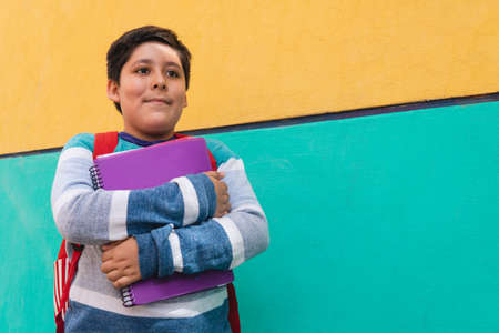 A Caucasian boy with black hair stands next to a yellow and green wall outside. The boy is in...