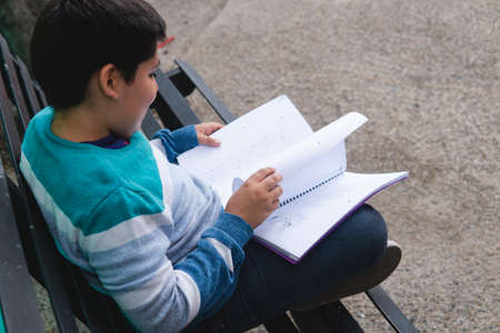 A black-haired boy in a striped sweater sitting on a park bench is reading a book and turning the...