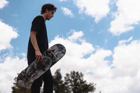 A skater with brown hair and a black shirt is on a ramp in the park with his skateboard in hand and is about to launch himself to perform some trick. The scene has a big sky in the backgroundの写真素材