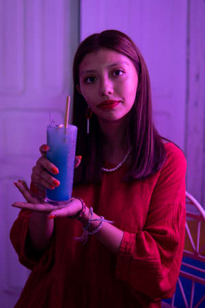 A young Hispanic woman wearing red and lipstick in a room with pink lighting with a blue colored drink in her hands. The girl is sitting in a bar with her drink. She models alongside the drink.の写真素材