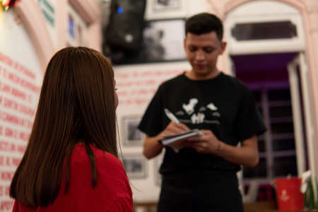 Hispanic girl with brown hair in a red dress in a nightclub ordering food and drinks from the waiter who takes notes in his notebook. Girl from behind looking at the waiter in the restaurant.の写真素材